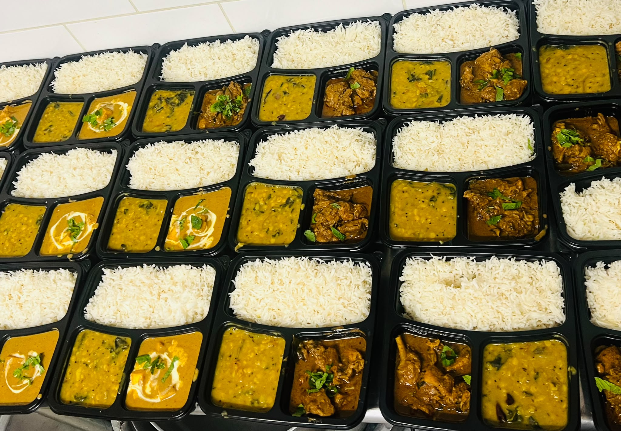 Rows of individual compartmentalized meal trays with rice, dal and curry, packed for office delivery